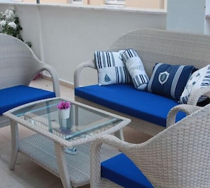 A wicker sofa with blue cushions and striped pillows, a glass table with a vase of flowers, and a white wall in the background.
