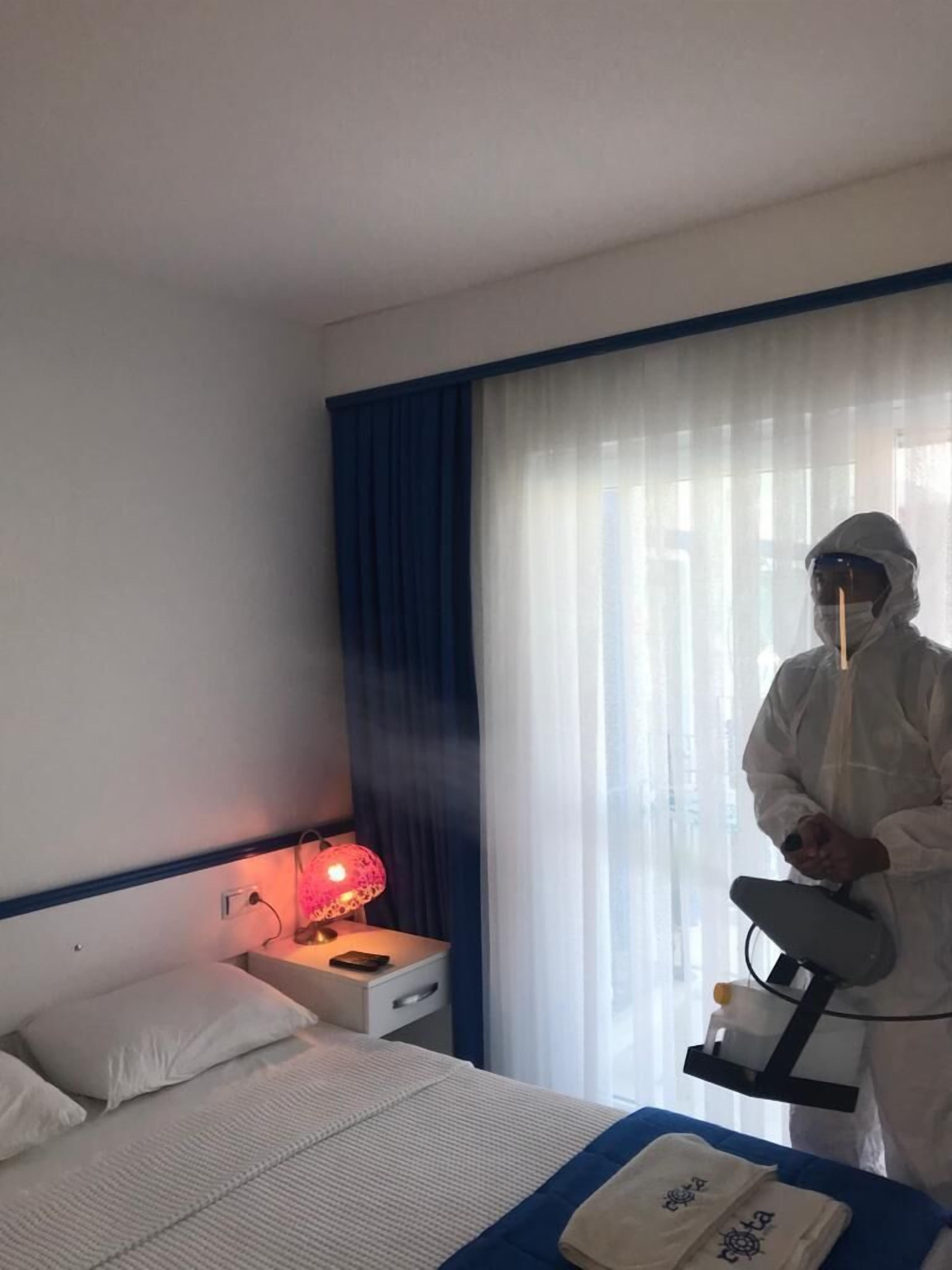 Une chambre d’hôtel avec un lit, une table de chevet avec une lampe et une personne portant un équipement de protection.