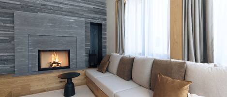 A modern living room with a fireplace, a wooden floor, a beige sofa with brown cushions, and a large window with curtains.