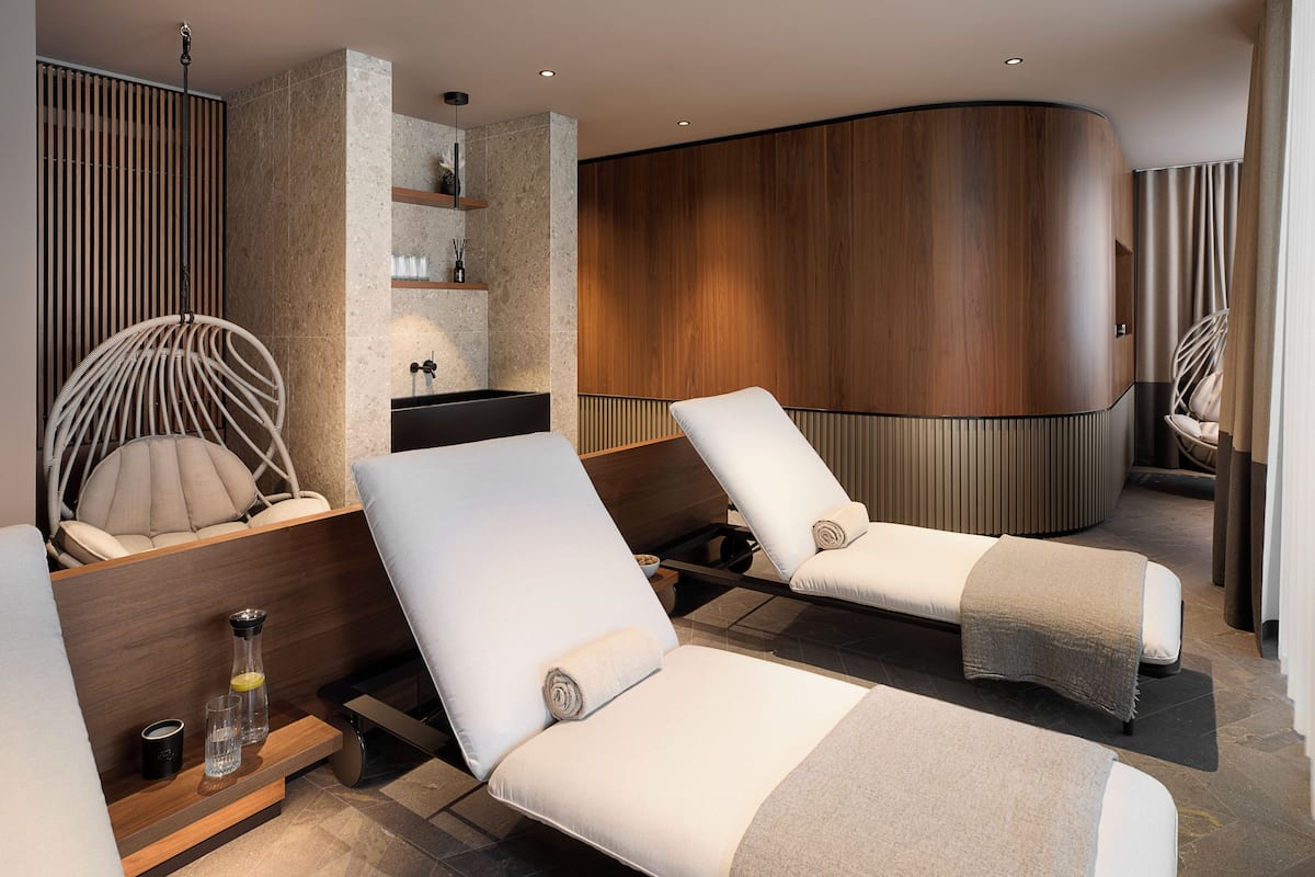 A modern spa room with two reclining chairs, a wooden panel wall, and a built-in shelf with toiletries.