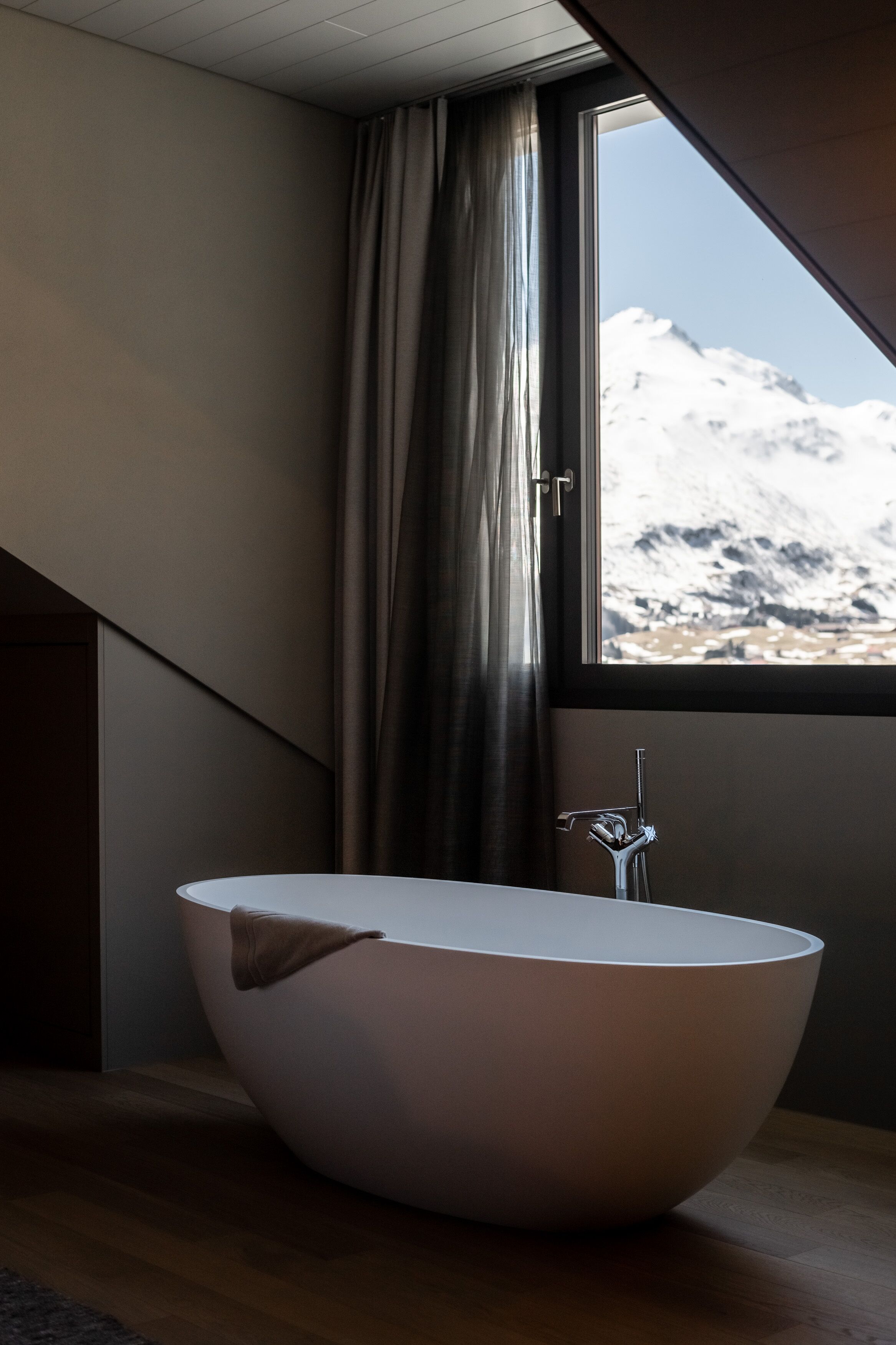 A modern bathroom with a freestanding bathtub, a view of a snowy mountain through a window, and a wooden floor.