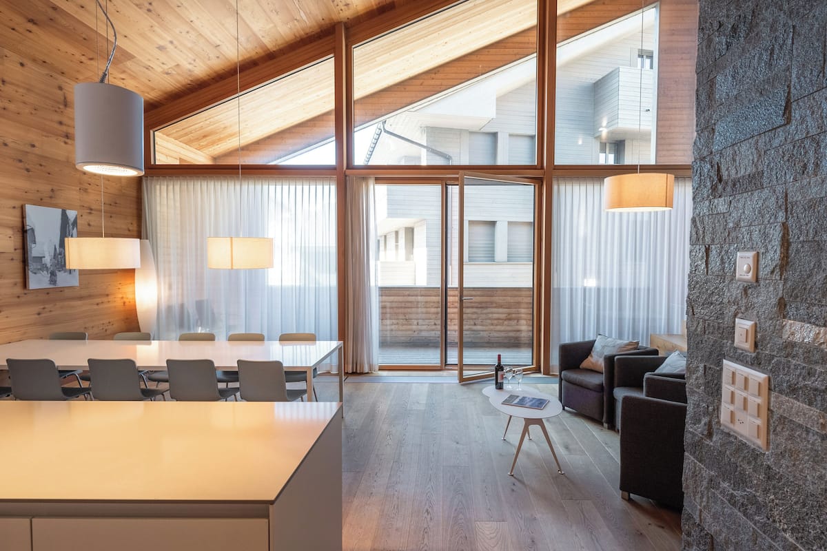 A modern interior with a wooden ceiling, large windows, a stone accent wall, a dining area, and a seating area with a small table and chairs.