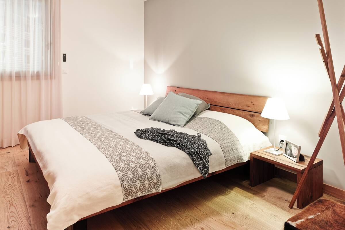 A bedroom with a wooden bed, bedside lamps, a bedside table with a photo, and a window with sheer curtains.