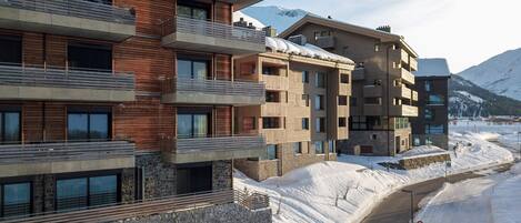 A modern multi-story building with balconies and large windows, surrounded by snow-covered ground and mountains in the background.