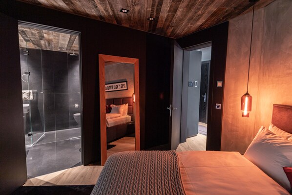 Una habitación de hotel moderna con una cama grande, cabina de ducha de vidrio, techo de madera y lámpara fijada a la pared.
