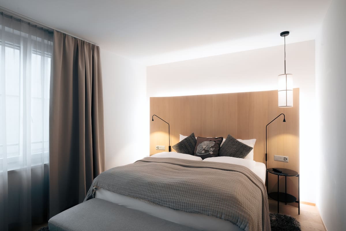 A modern bedroom with a wooden headboard, a bedside table with a lamp, and a large window with curtains.