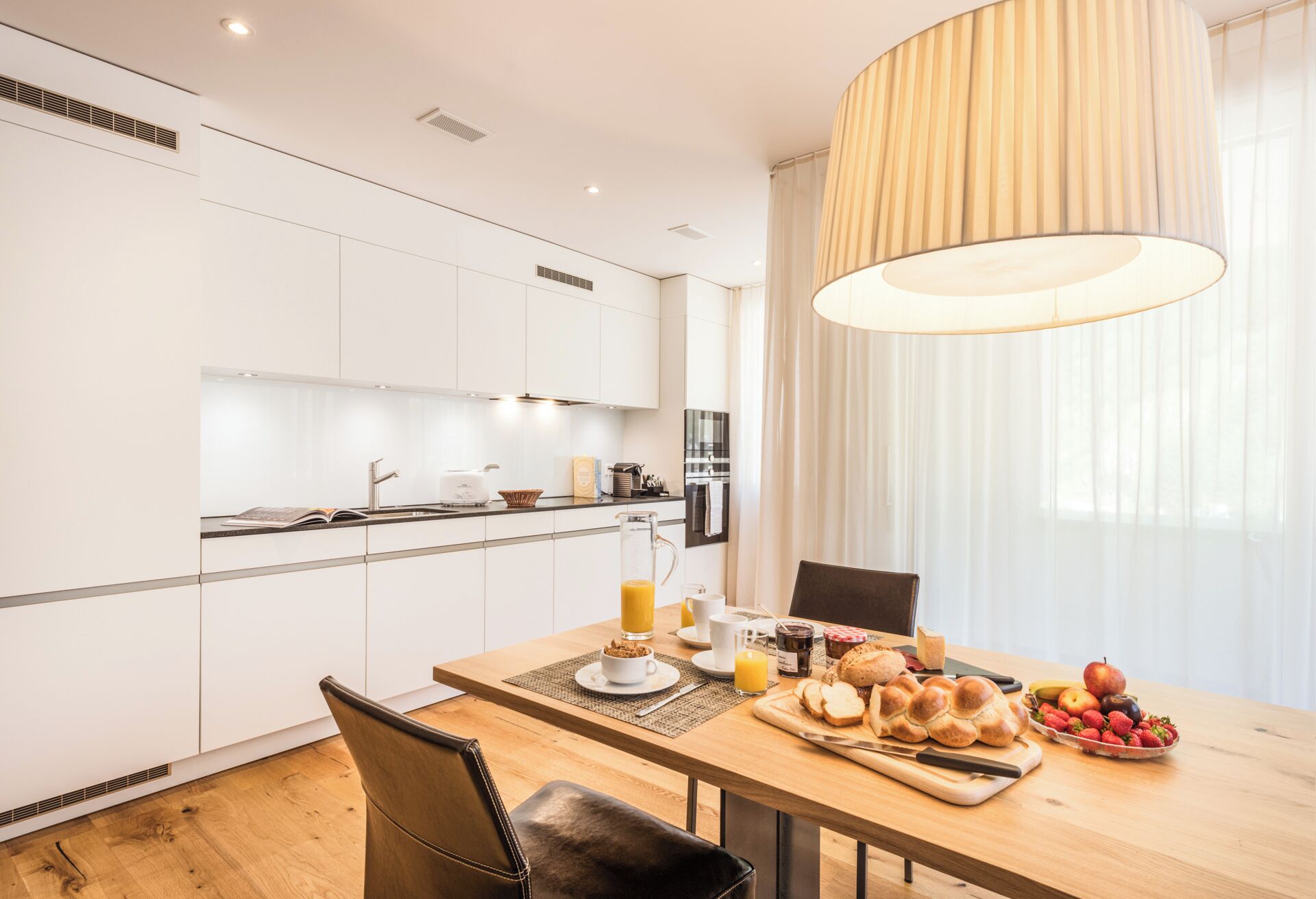 A modern kitchen with white cabinets, a dining table with a placemat, and a breakfast setting with bread, fruit, and juice.