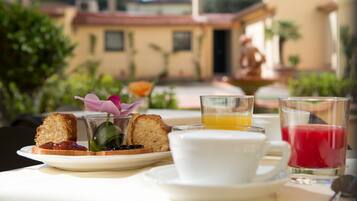 Un desayuno con tostadas, mermelada, un pequeño florero con una flor, una taza y vasos de jugo de naranja y jugo de cereza sobre una mesa.
