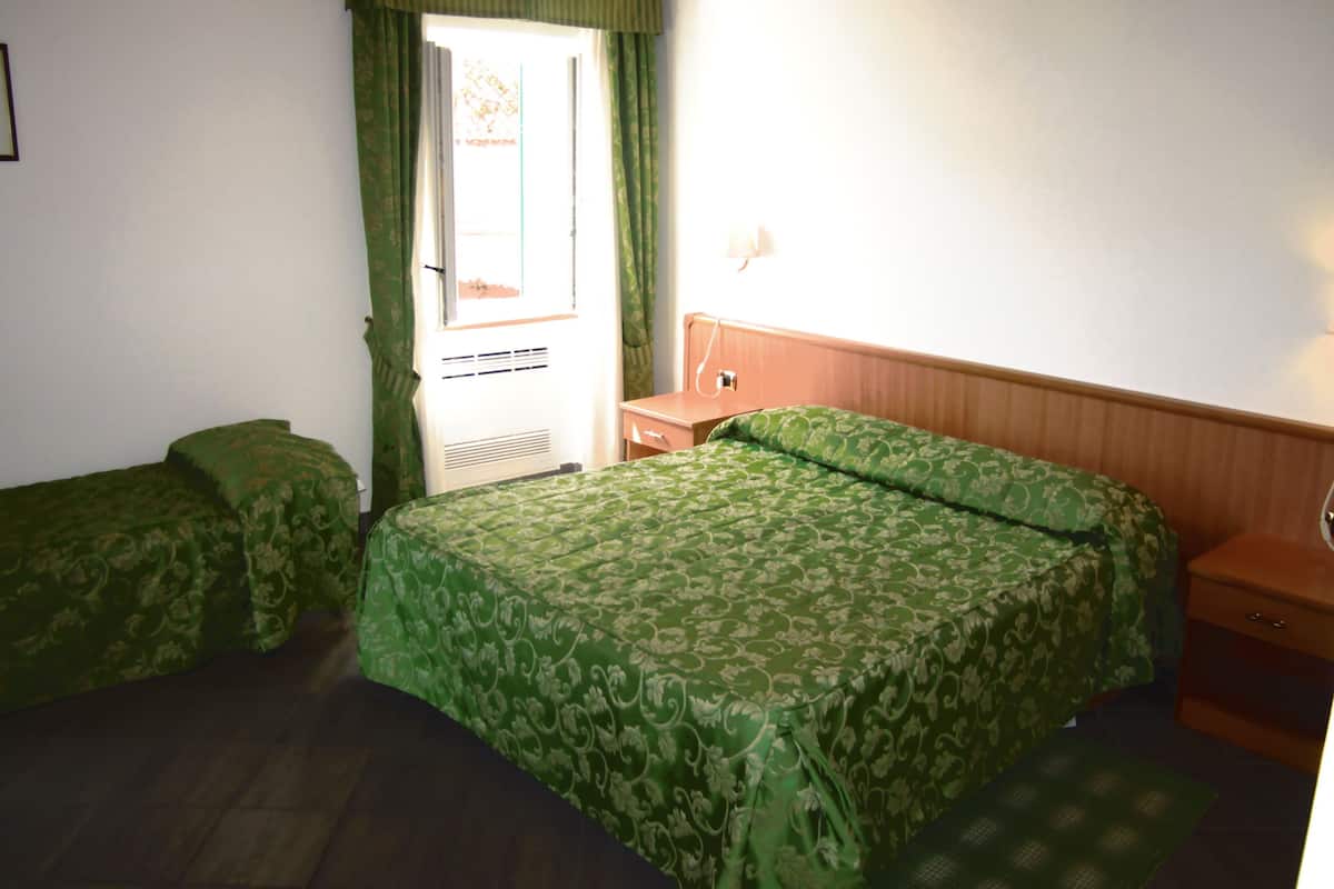 A room with two beds, a wooden nightstand, a wall-mounted lamp, and a window with green curtains.
