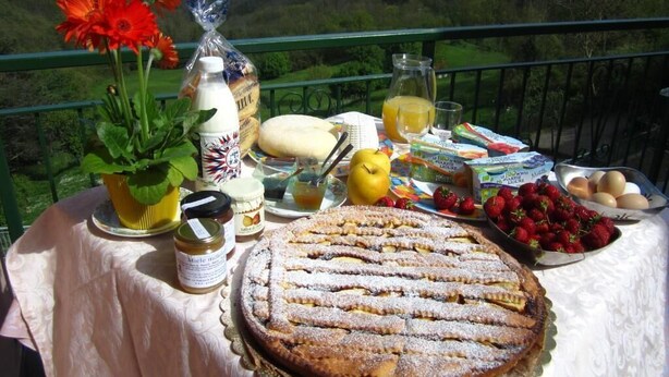 Una mesa con un postre, fruta y aderezos en un balcón con vista a la montaña.
