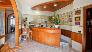 Una zona de recepción con un mostrador de madera, una computadora y una planta. Hay cuadros enmarcados en la pared y una pequeña área de descanso con una silla.