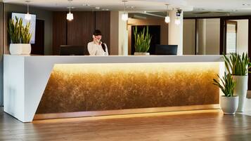 Una reception dal design moderno, con una texture simile al marmo e una luce calda. Ai lati dell'area reception ci sono piante in vaso, e una receptionist è in piedi dietro di essa.