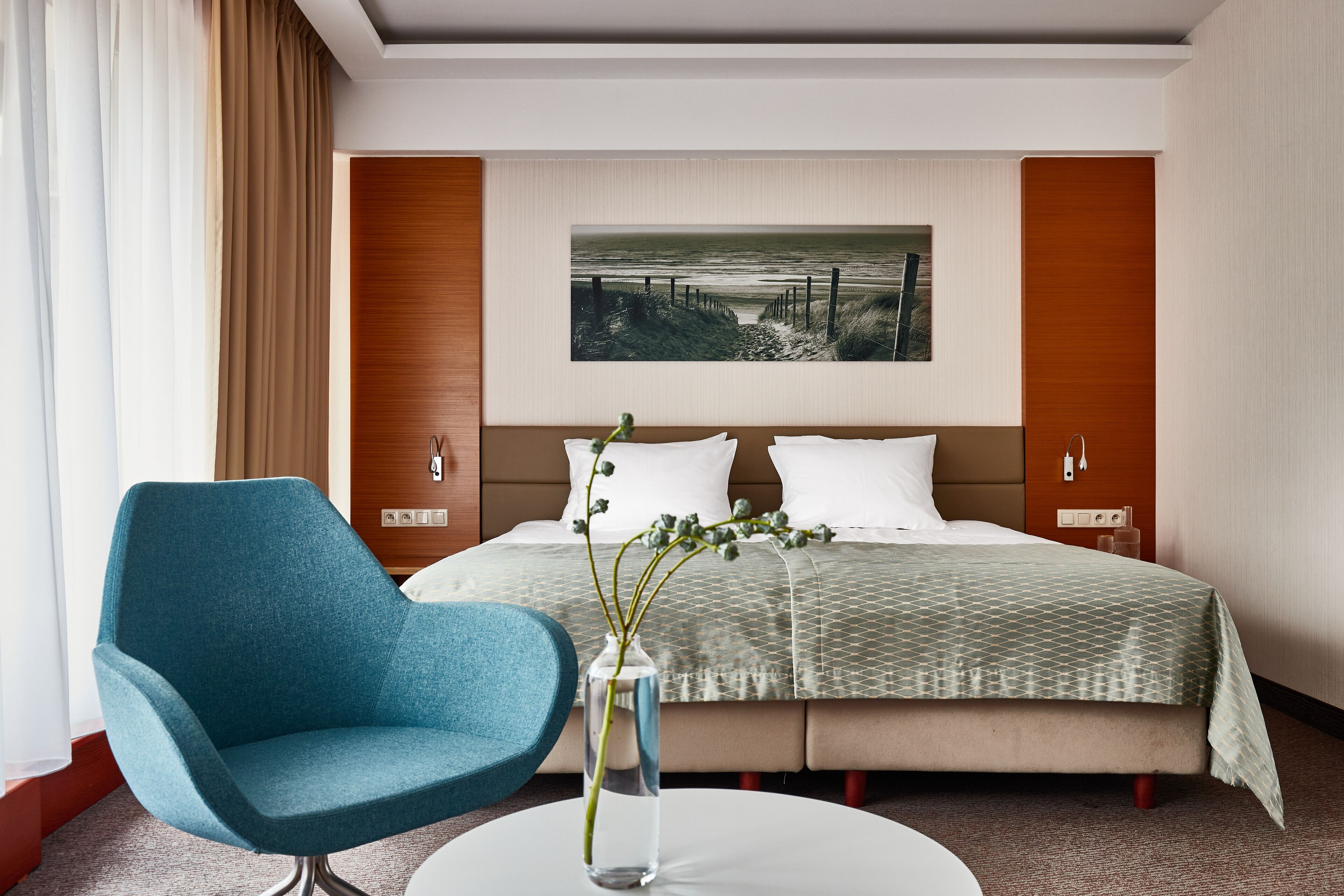 Una habitación de hotel con una cama, un sillón azul, un jarrón con una planta y un cuadro en la pared.