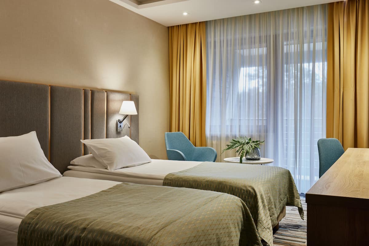 Habitación de hotel con dos camas, un escritorio, una silla, una lámpara y un ventanal con cortinas.