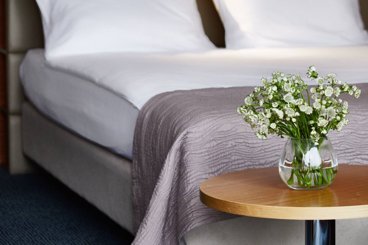 Habitación de hotel con cama, almohadas blancas, una mesita con un florero y un cuadro en la pared.