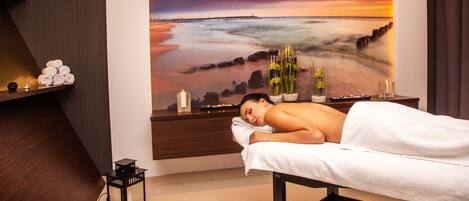 A massage room with a large wall mural of a beach sunset, a wooden bench, and a small shelf with candles and flowers.