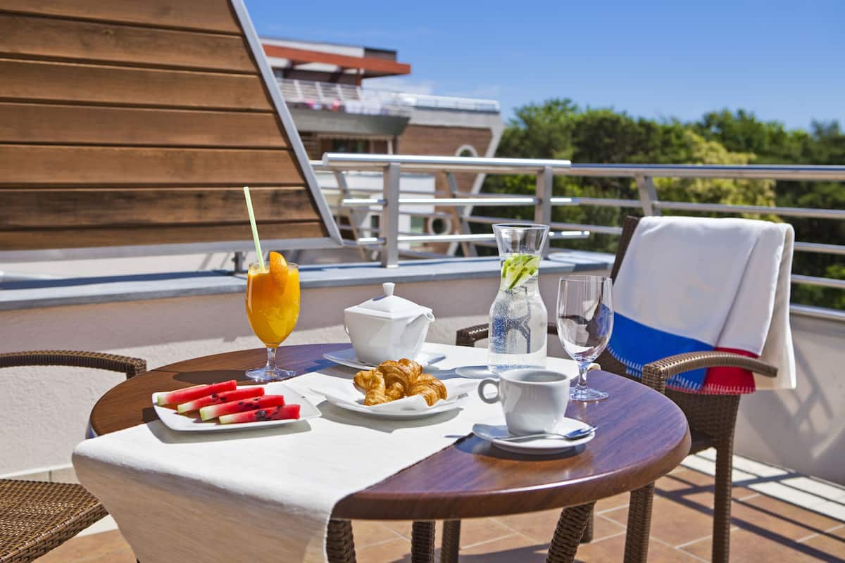 Una mesa dispuesta con cruasanes, rebanadas de sandía, un juego de té, un vaso de zumo de naranja y una botella de agua en un balcón con vistas a un edificio y árboles.