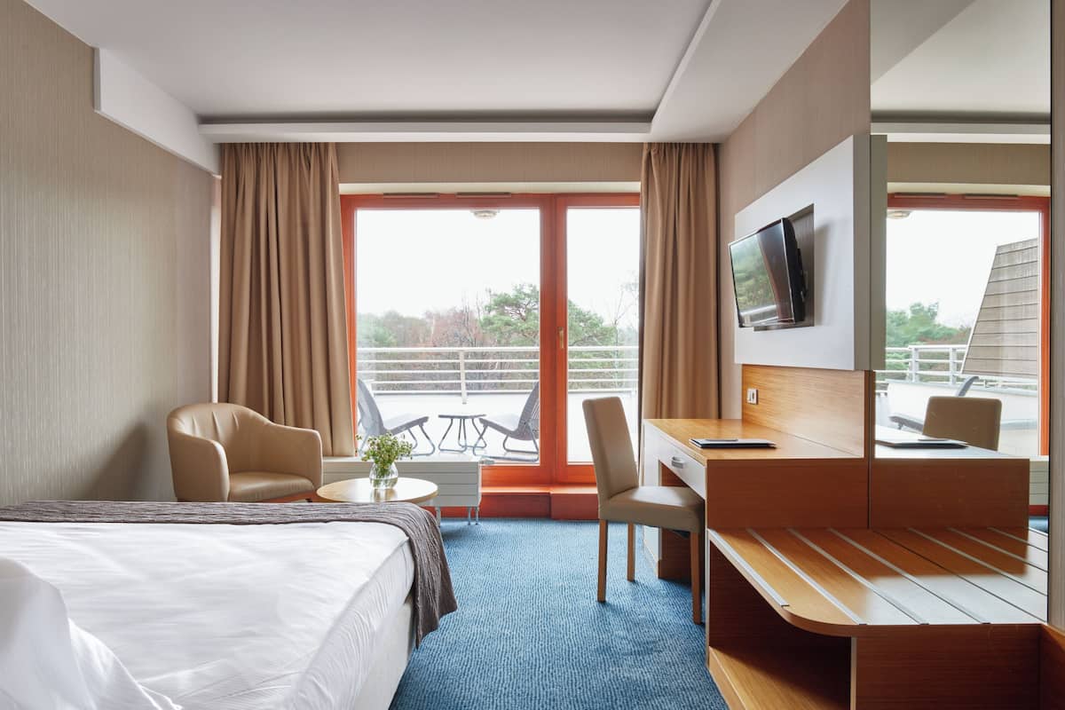 Habitación de hotel con cama, escritorio, silla, televisor y balcón con mobiliario exterior.