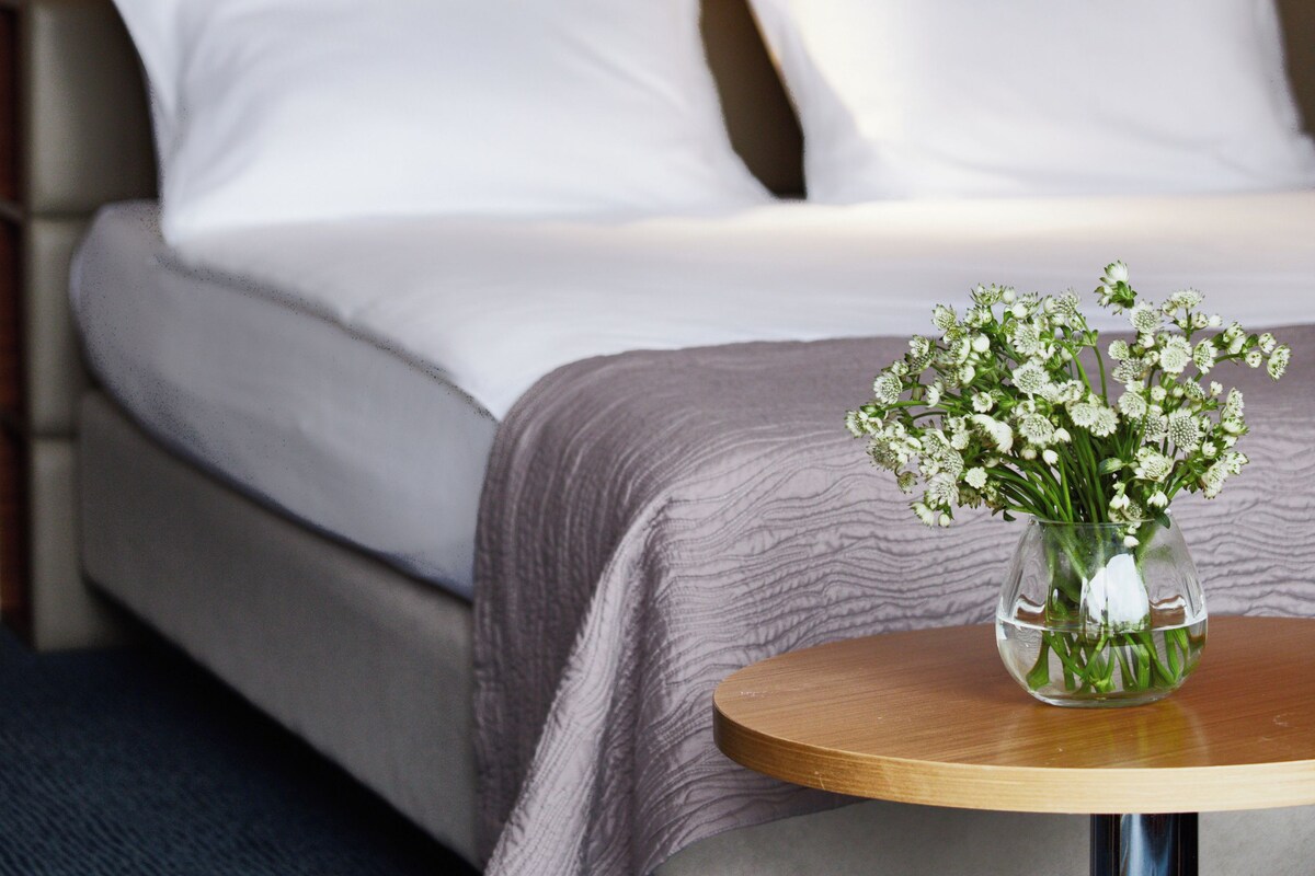 A hotel room with a bed, white pillows, a small table with a vase of flowers, and a framed picture on the wall.