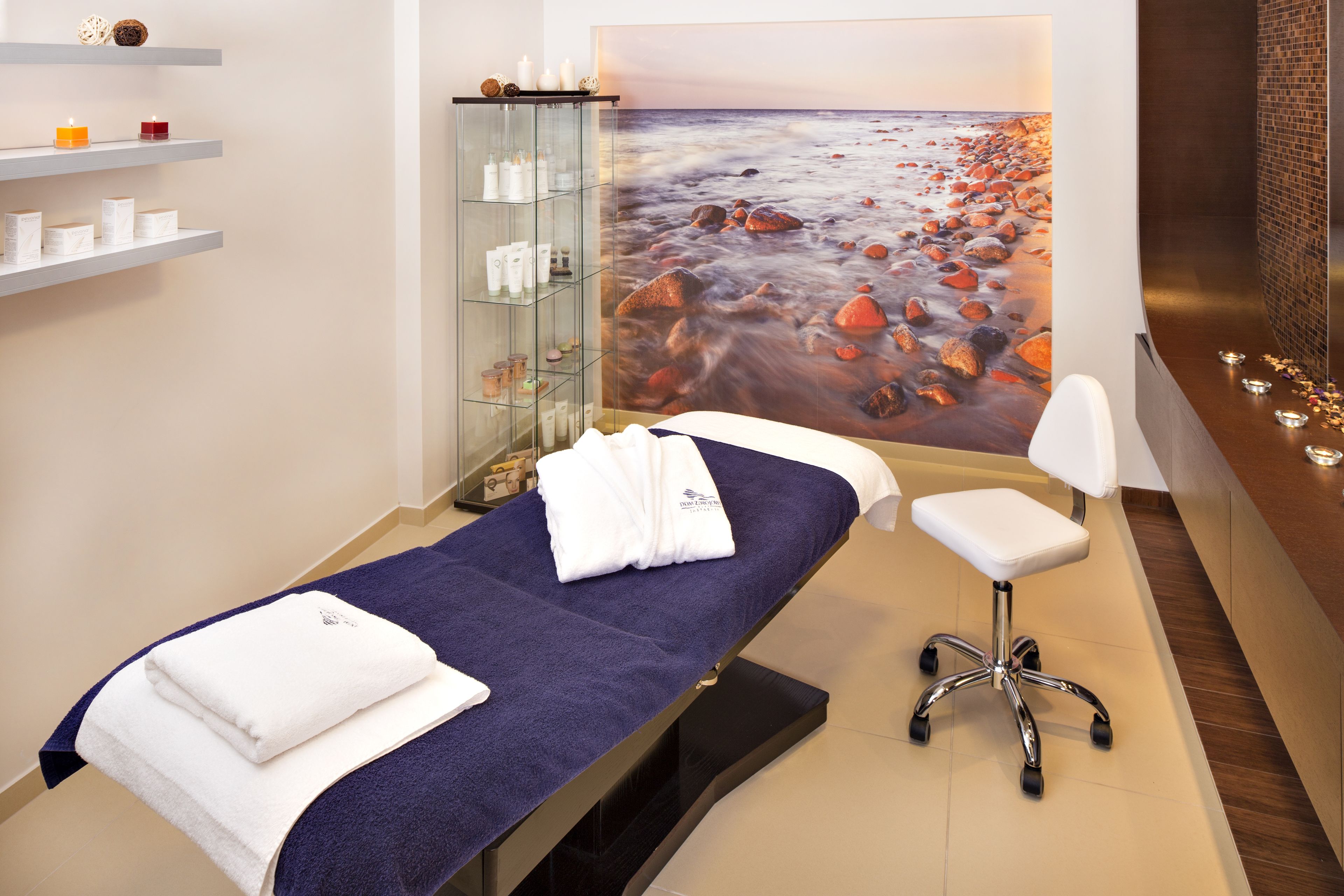 A spa room with a massage table, a white chair, and a shelf with candles and toiletries.