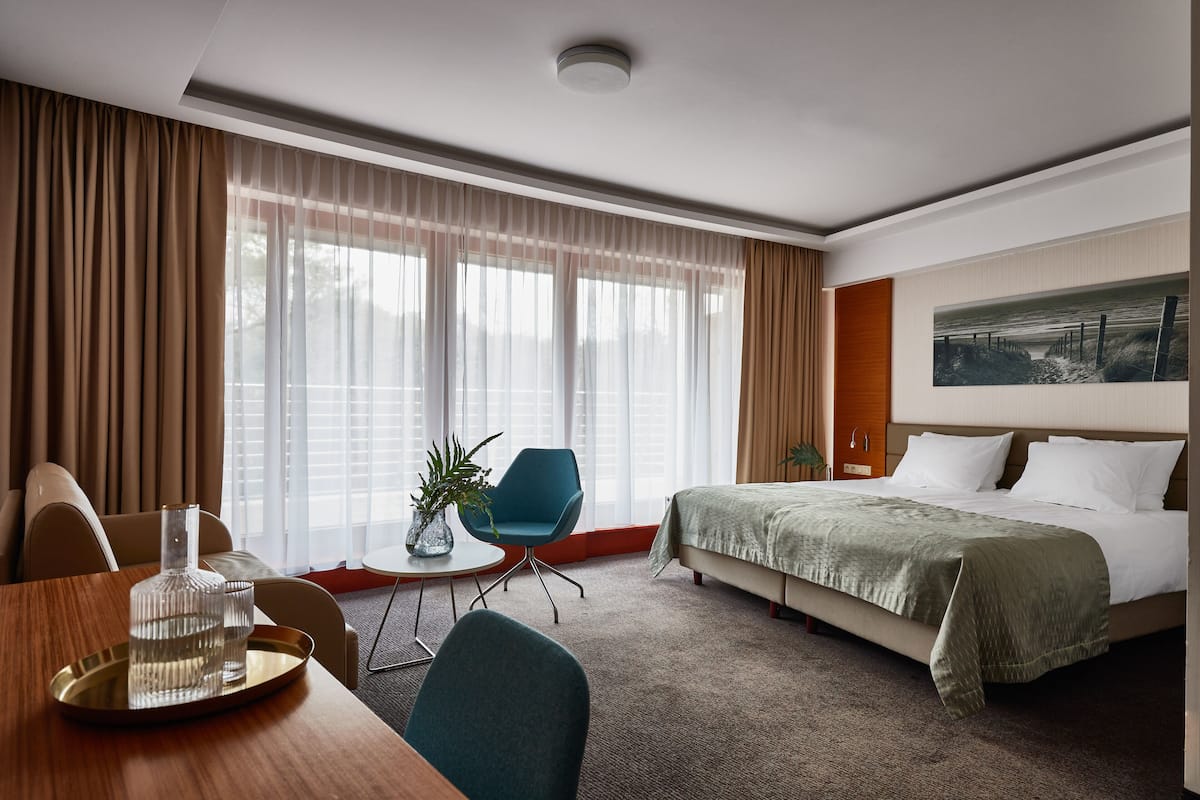 Una habitación de hotel con cama, una mesa de comedor con sillas, una mesita auxiliar y un ventanal con cortinas.