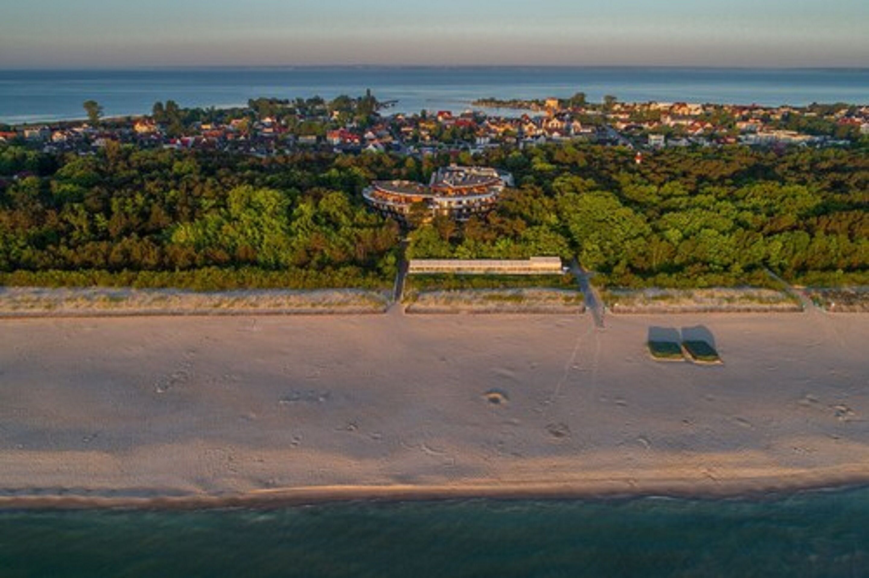 An aerial view of a coastal area with a large building surrounded by dense greenery, a sandy beach, and a clear view of the sea.