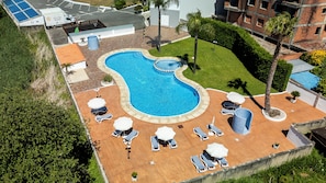 Une piscine entourée de verdure, avec des chaises longues et des parasols, ainsi qu’un espace de stationnement.