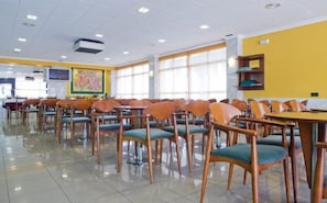 Un vaste espace de restauration intérieur, avec des rangées de chaises et de tables en bois, de grandes fenêtres et un projecteur fixé au plafond.