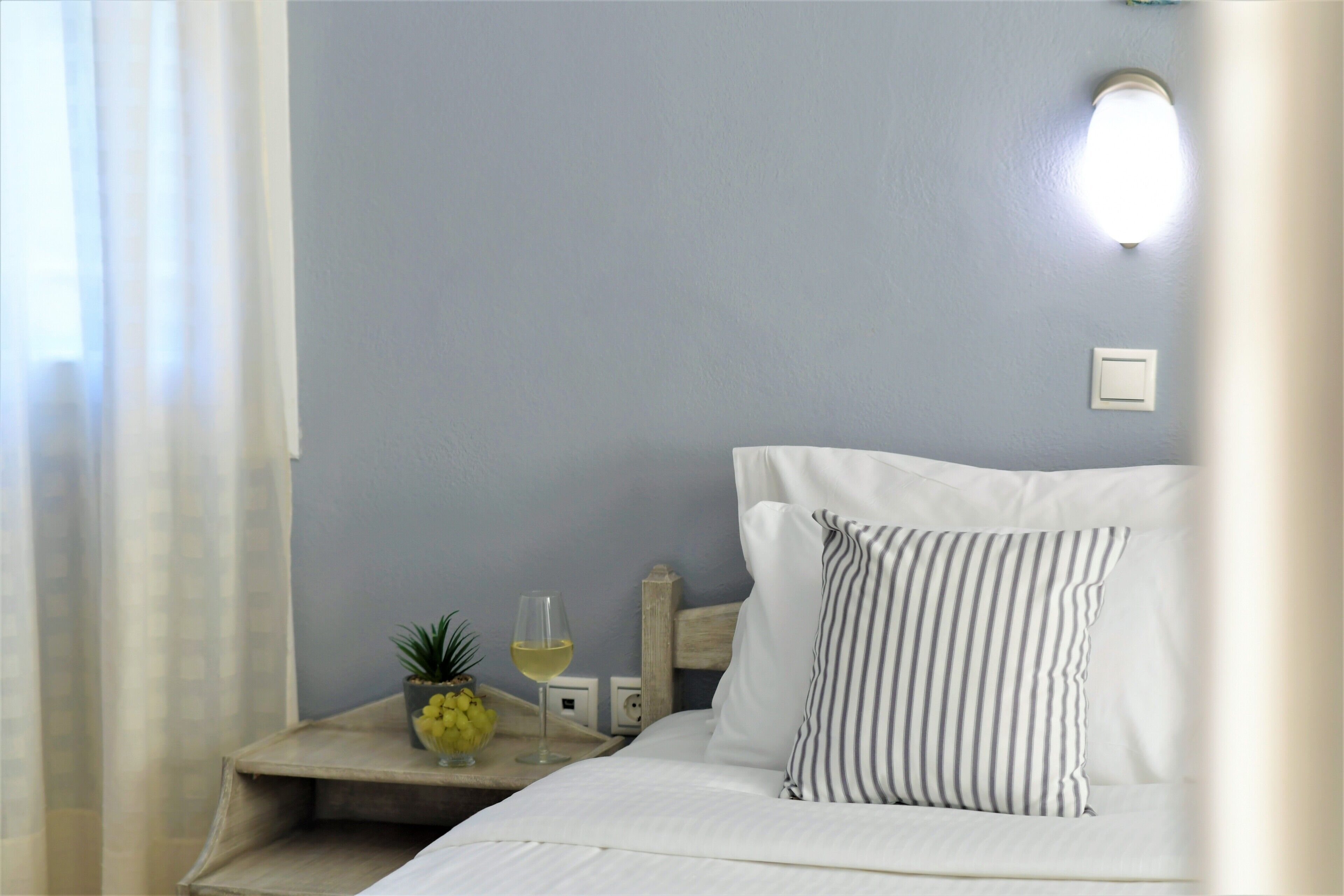 A neatly made bed with white linens and striped pillows, a small bedside table with a pineapple and a glass of white wine, and a wall-mounted light fixture.