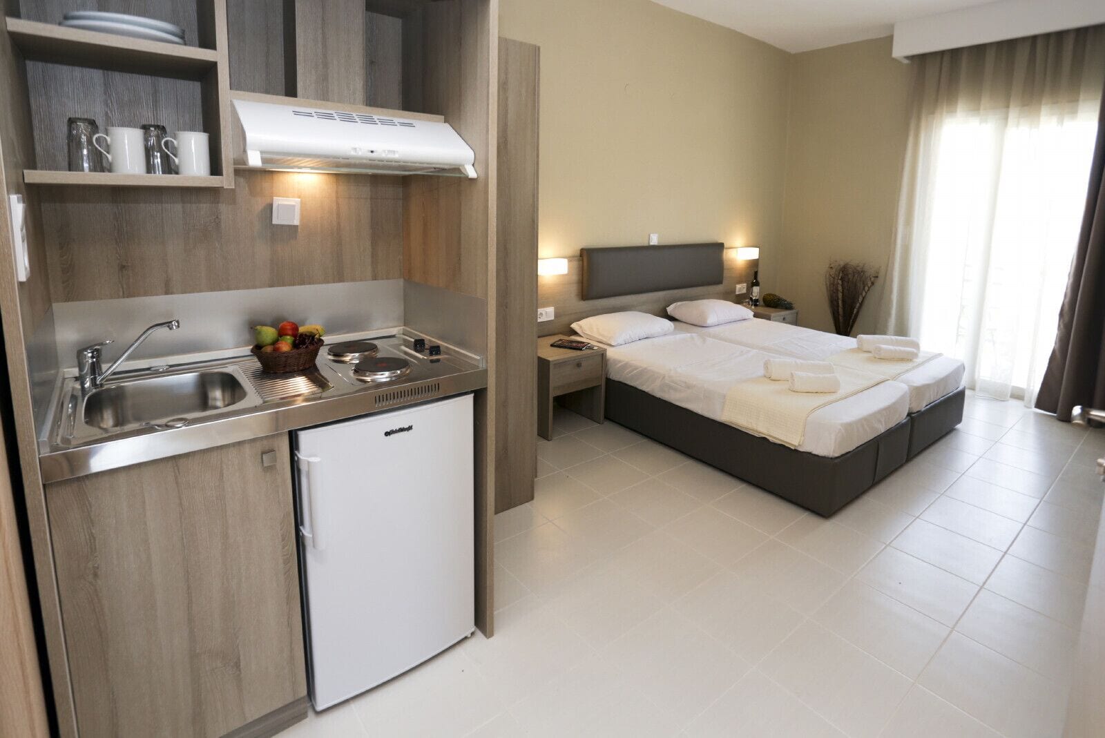 Una habitación de hotel pequeña con kitchenette, una cama con dos almohadas y una mesita de noche con lámpara.