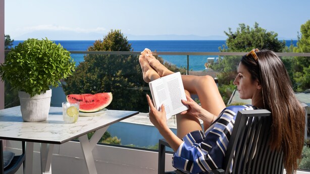 Una persona leyendo un libro en un balcón con vista al mar, una planta en maceta y una rebanada de sandía sobre la mesa.