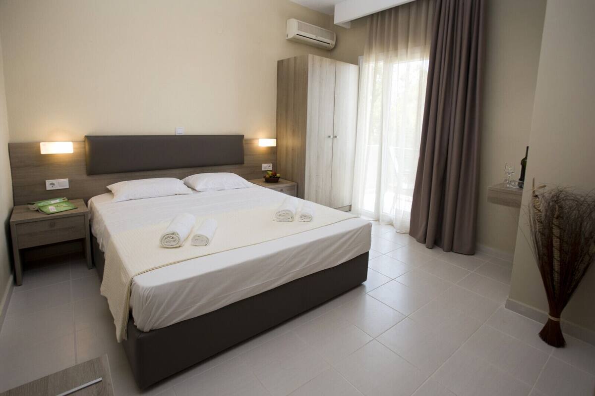 Une chambre d’hôtel moderne avec un grand lit, deux tables de chevet, une petite table, un climatiseur fixé au mur et une fenêtre avec des rideaux.