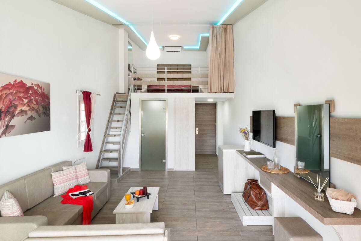 Un salon moderne avec un canapé, une table basse et un téléviseur posé sur une console en bois. Un escalier mène à un lit superposé situé à l’étage.