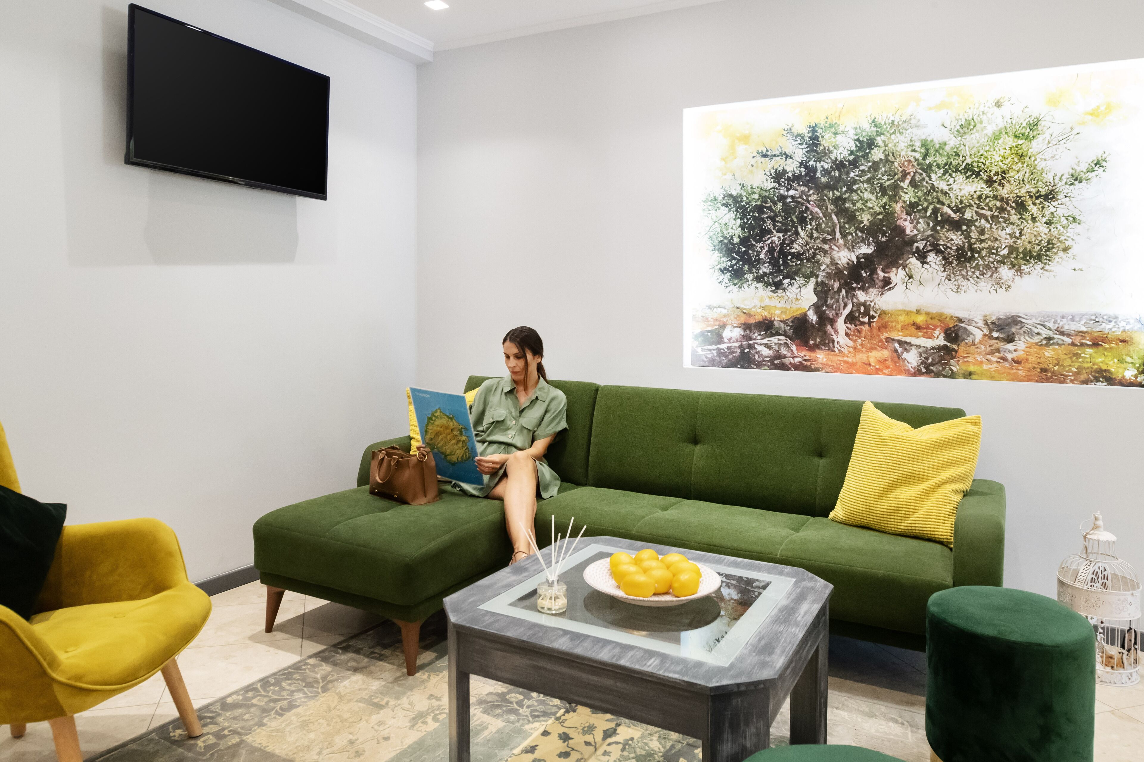 A living room with a green sofa, a yellow armchair, a coffee table with a bowl of lemons, and a painting of a tree on the wall.
