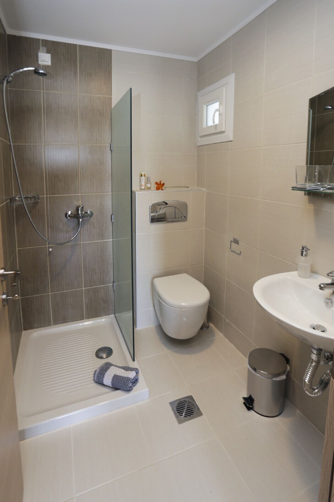 A bathroom with a shower, toilet, sink, and a small trash bin.