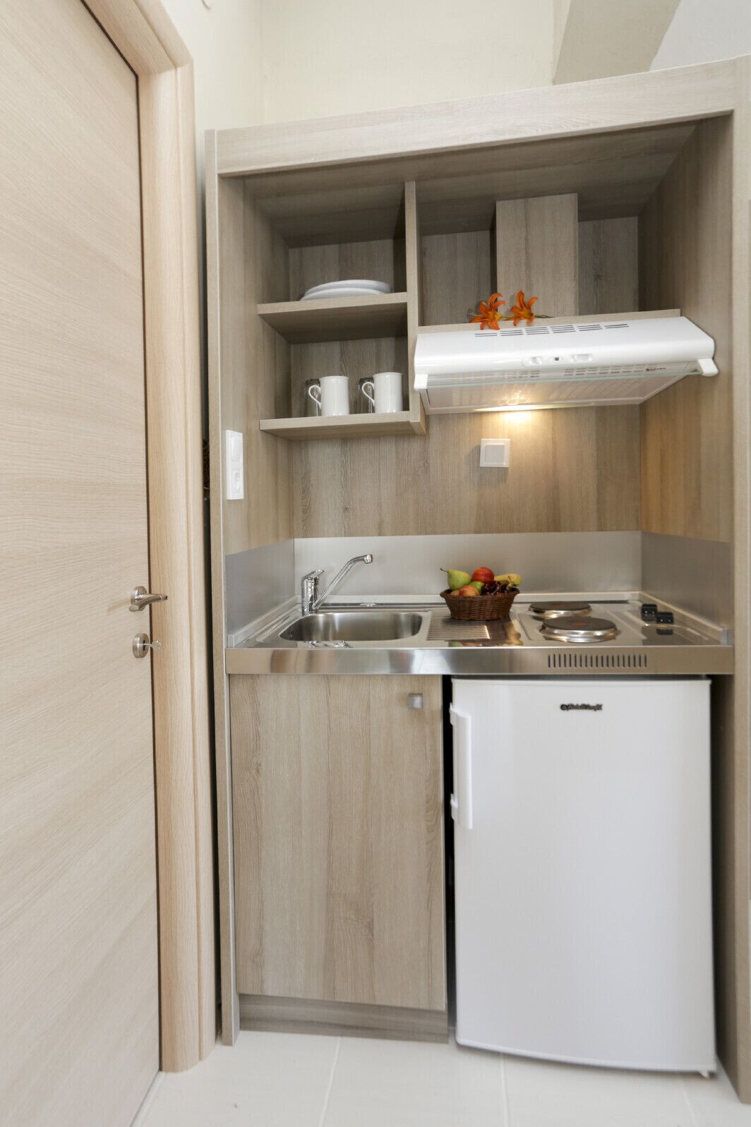 Cocina compacta con pileta integrada, un refrigerador pequeño y un microondas. Cuenta con alacenas de madera y un estante con tazas y una cesta de frutas.