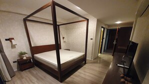 A four-poster bed with a wooden frame and a mattress, a small bedside table with a plant, a television on a stand, and a wooden floor. - Big Tree Old Town Hotel (Chiang Mai)