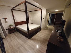 A four-poster bed with a wooden frame and a mattress, a small bedside table with a plant, a television on a stand, and a wooden floor. - Big Tree Old Town Hotel (Chiang Mai)