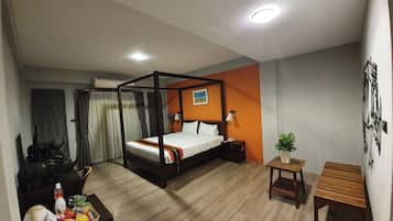 Habitación de hotel con una cama con dosel, una mesita de noche con lámpara, una pequeña mesa de madera, una planta en maceta y un escritorio con silla.