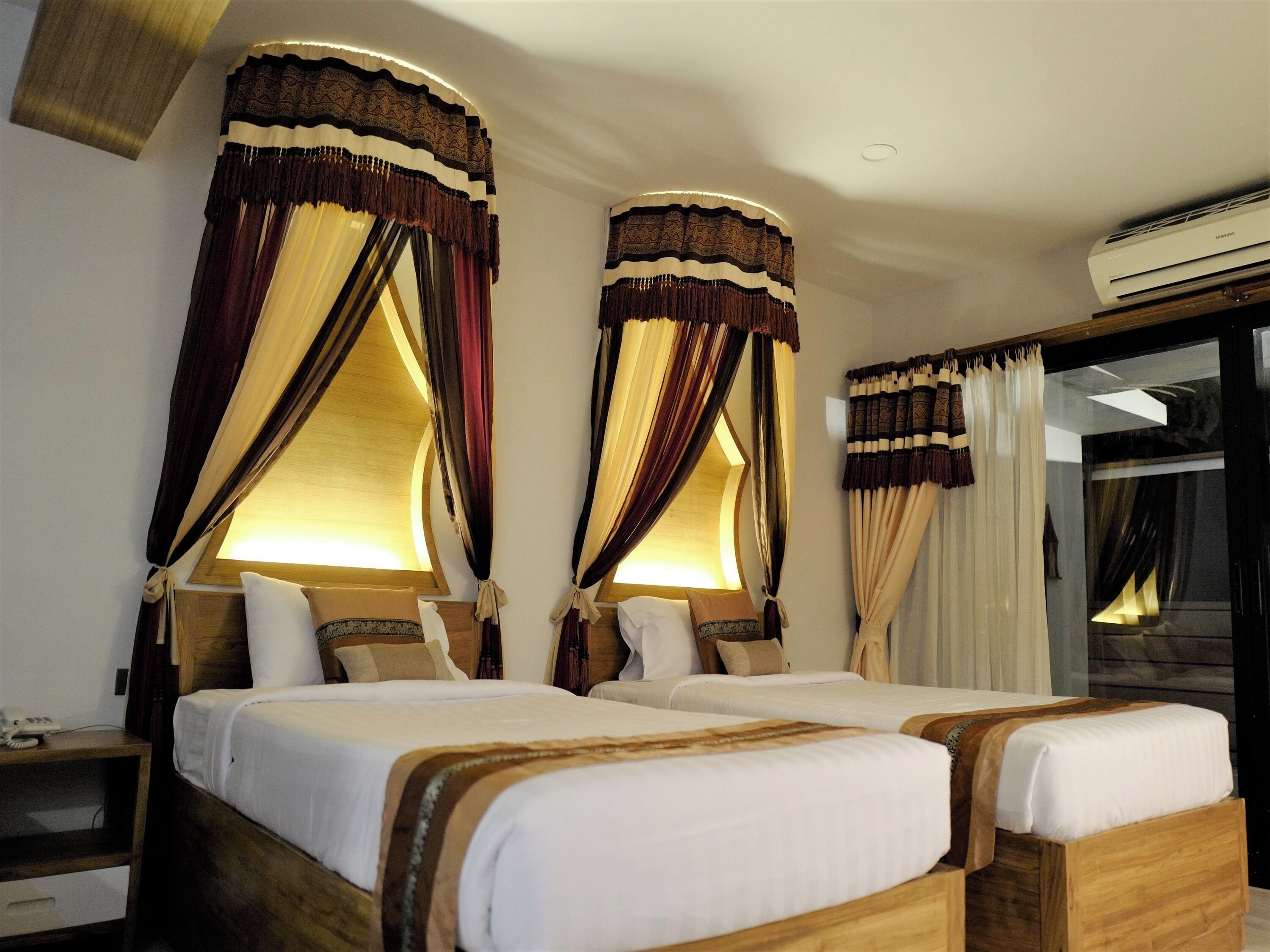A hotel room with two beds, a wooden headboard, a bedside table with a lamp, and a large window with curtains.