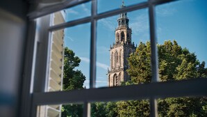 A view through a window of a tall, ornate tower with intricate architectural details and a clock, surrounded by lush green trees. - Hotel Prinsenhof (Groningen)