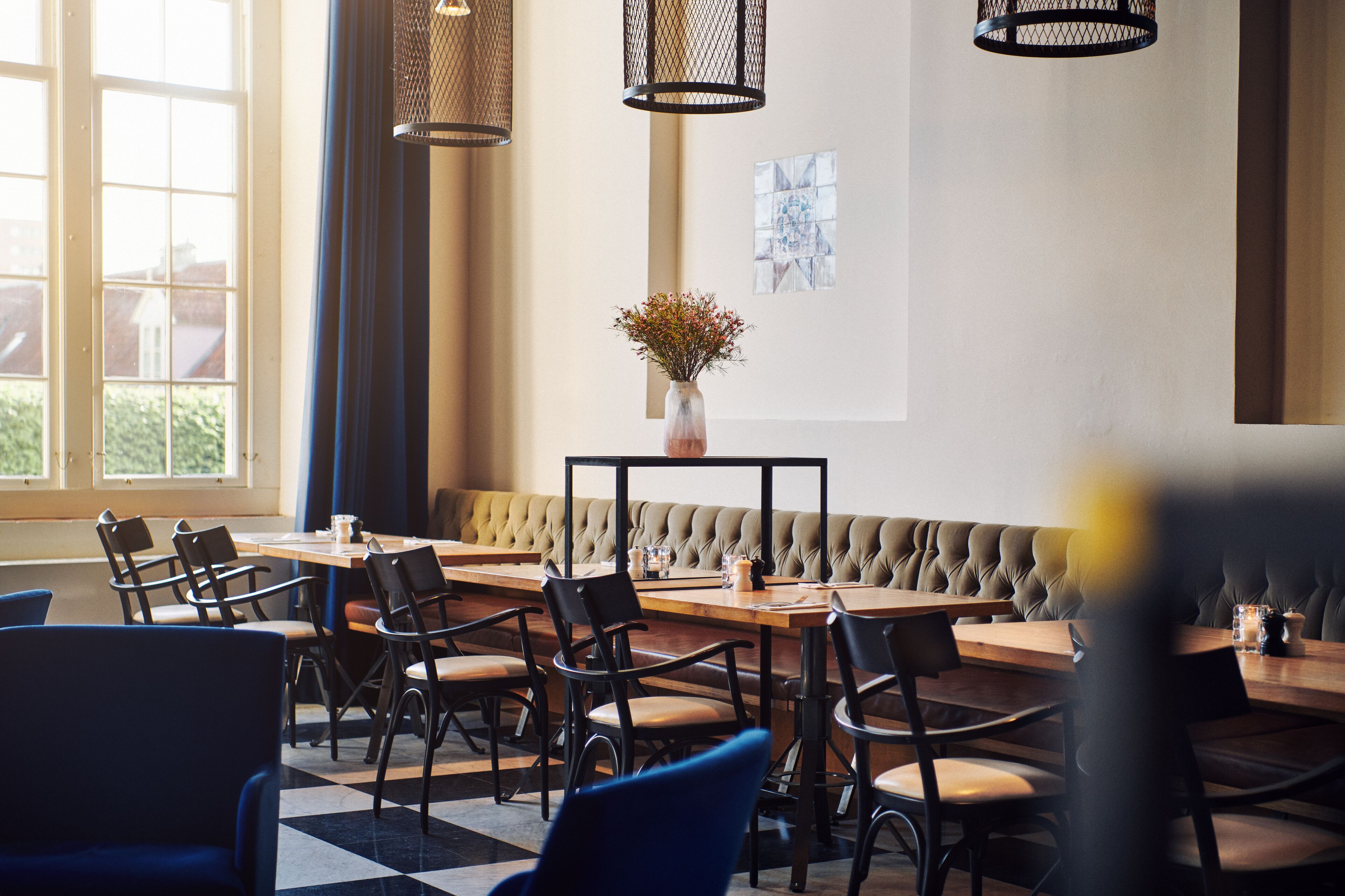 A restaurant with a checkered floor, wooden tables, and black chairs. There is a cushioned bench and a vase with flowers on a table.