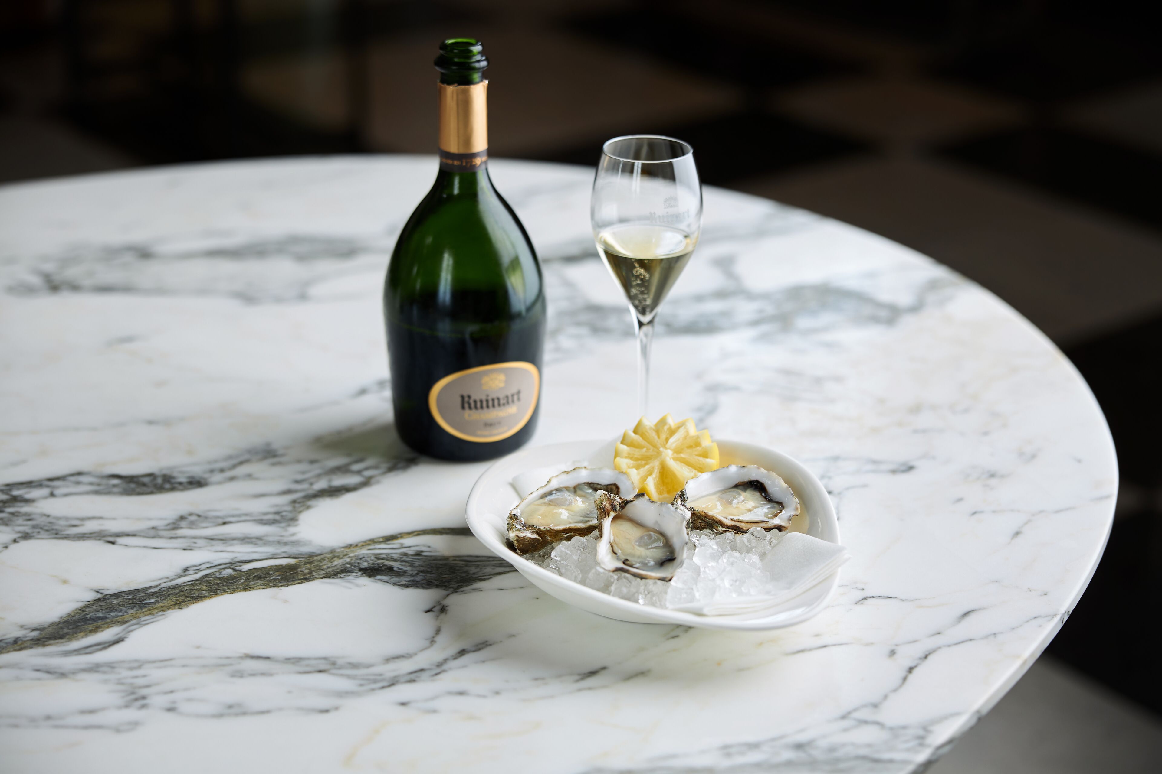 A bottle of Ruinart wine, a glass of white wine, and a plate of oysters on ice on a marble table.