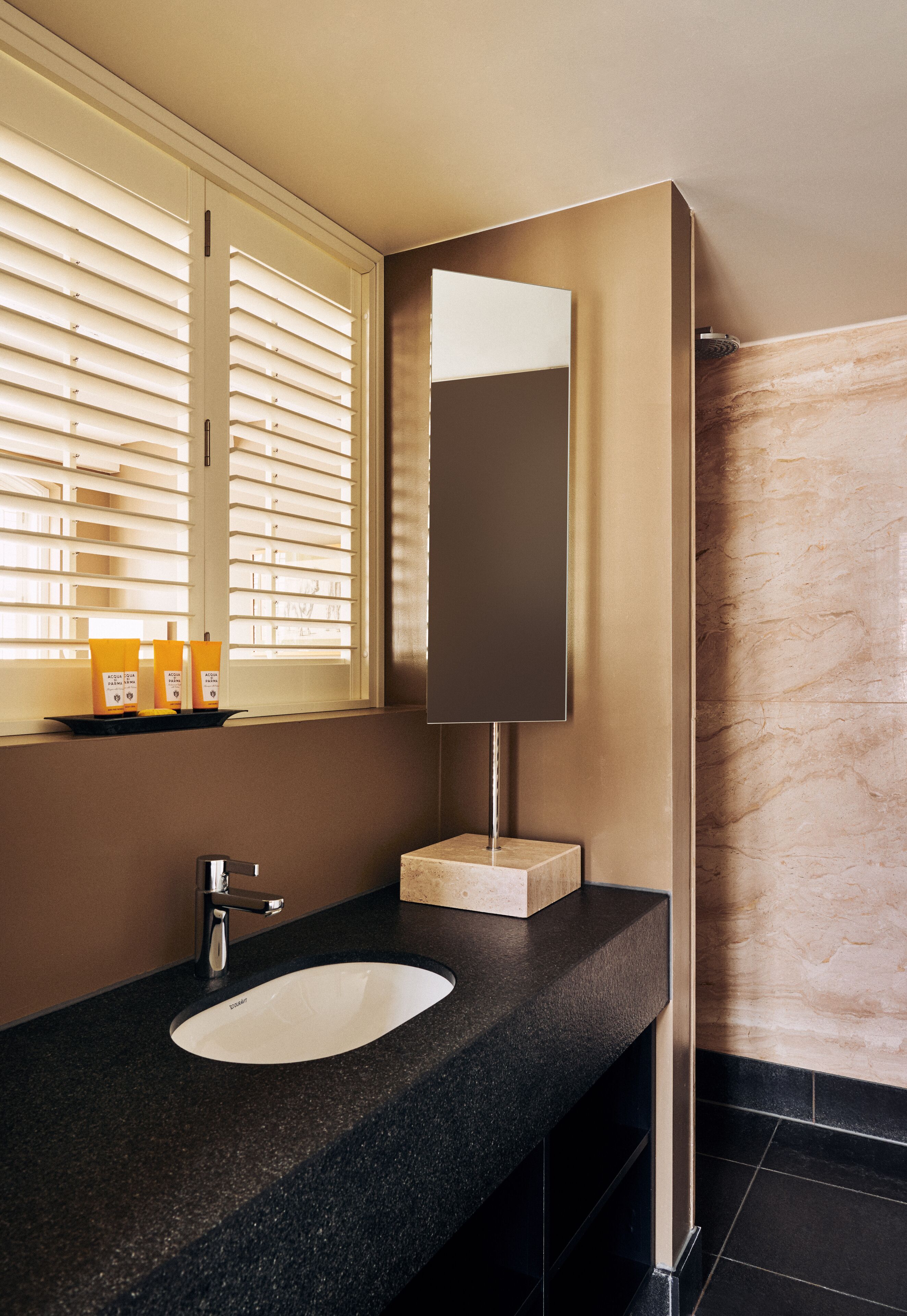 Un baño moderno con encimera de granito negro, lavabo blanco, espejo y ventana con persianas.
