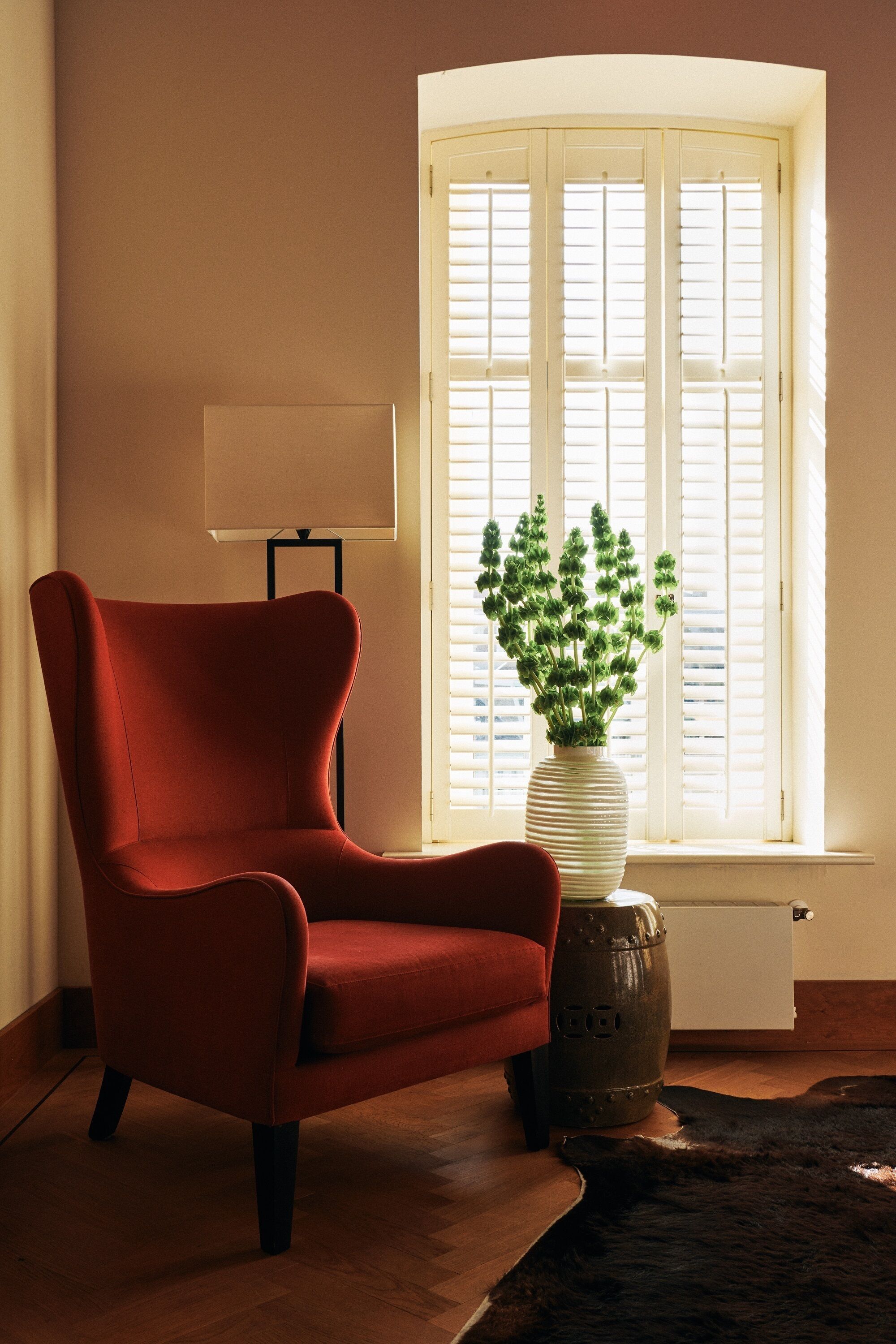 A room with a red armchair, a white radiator, a window with blinds, a lamp, and a vase with green plants.