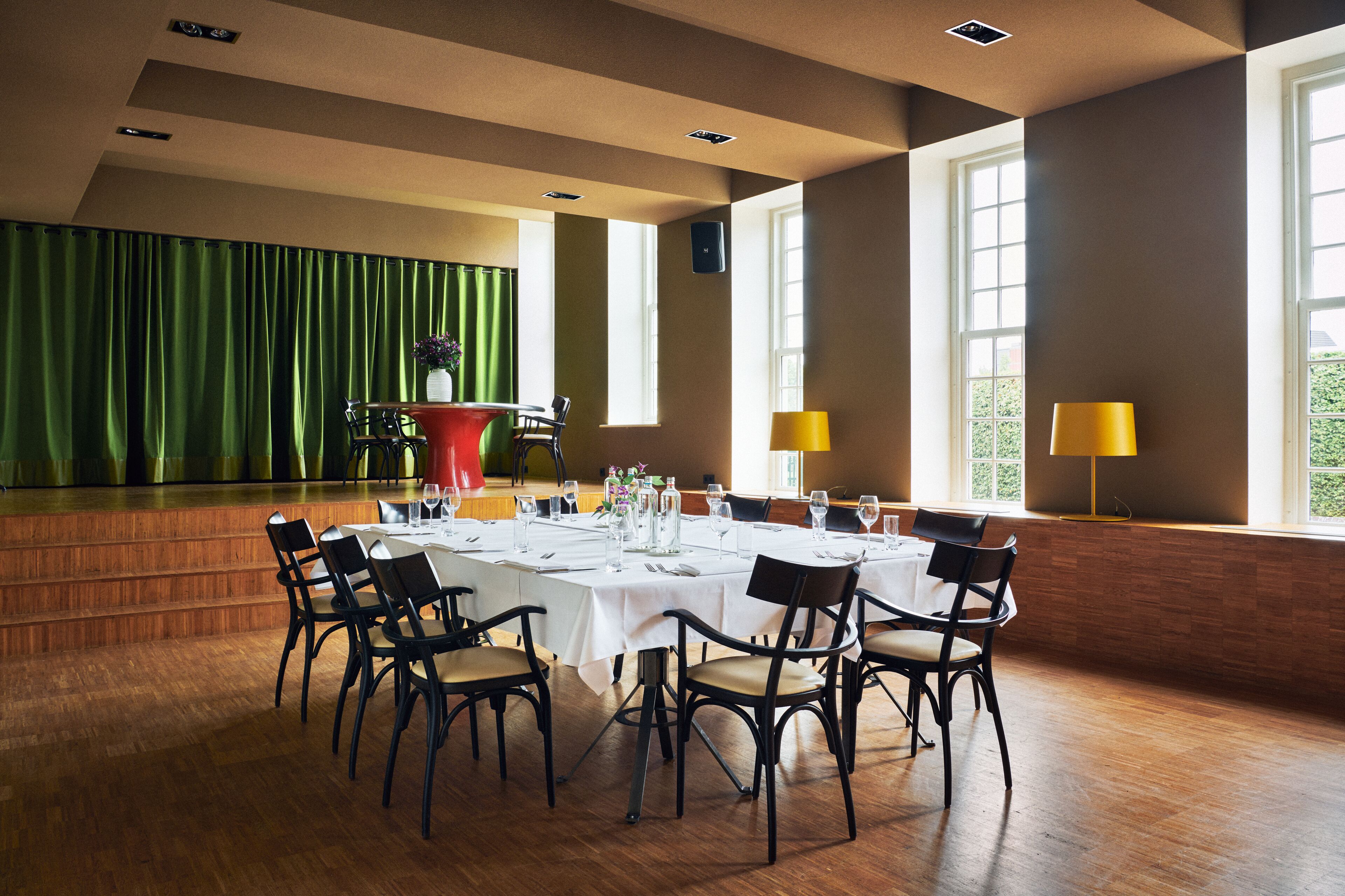 A dining area with a long table set for a meal, surrounded by chairs, with a stage and curtains in the background.