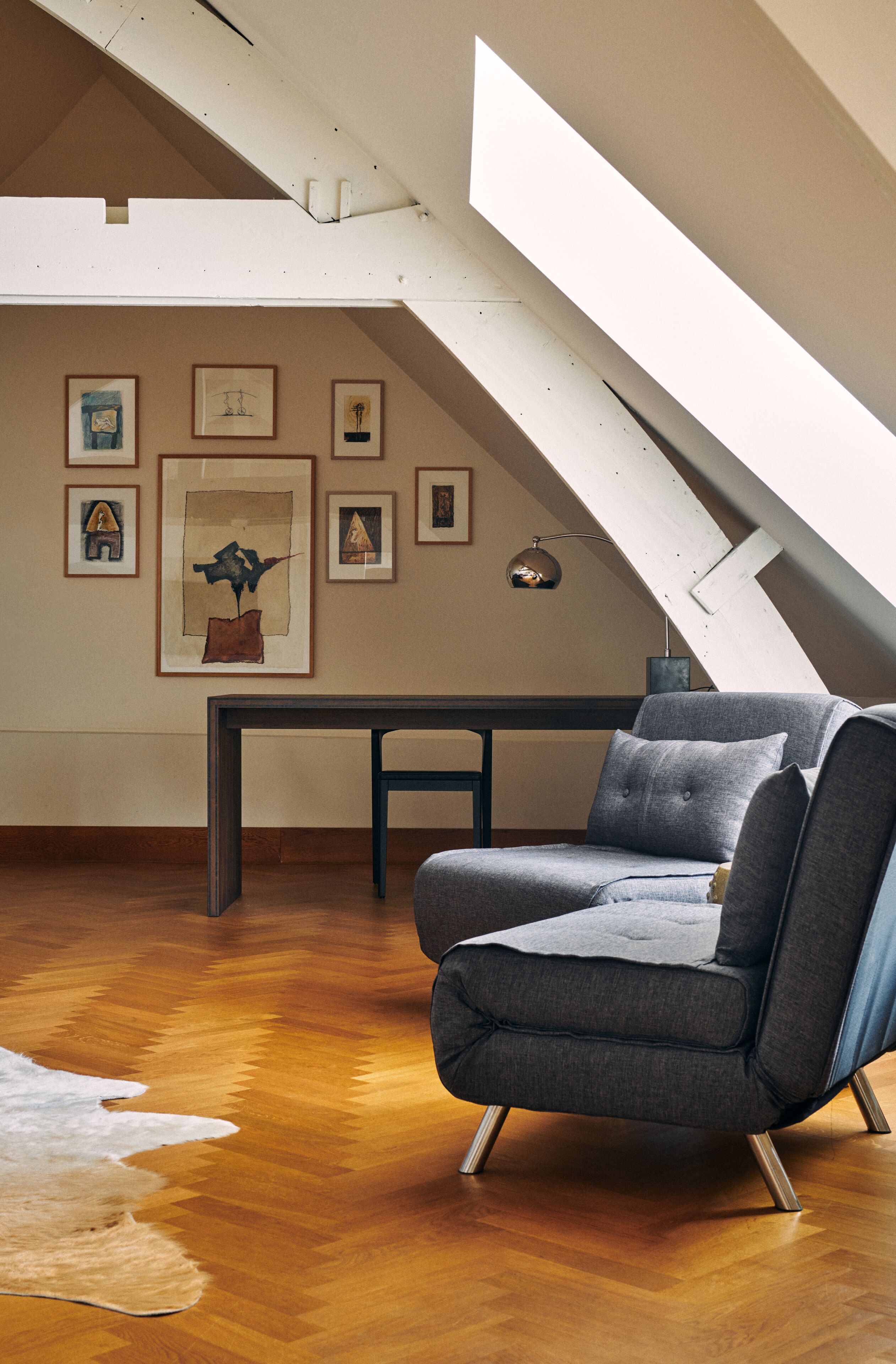 Un habitación estilo loft con techo inclinado, piso de madera, un sofá gris, una pequeña mesa y cuadros en las paredes.