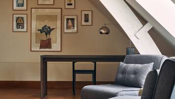 Un habitación estilo loft con techo inclinado, piso de madera, un sofá gris, una pequeña mesa y cuadros en las paredes.