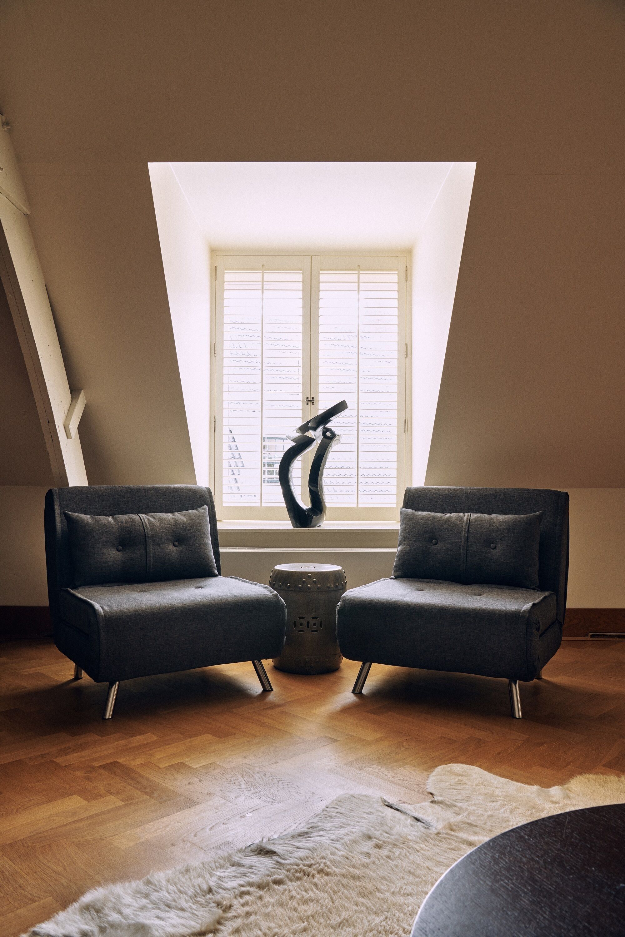 A room with a sloped ceiling, two dark armchairs, a small round table, and a window with blinds.