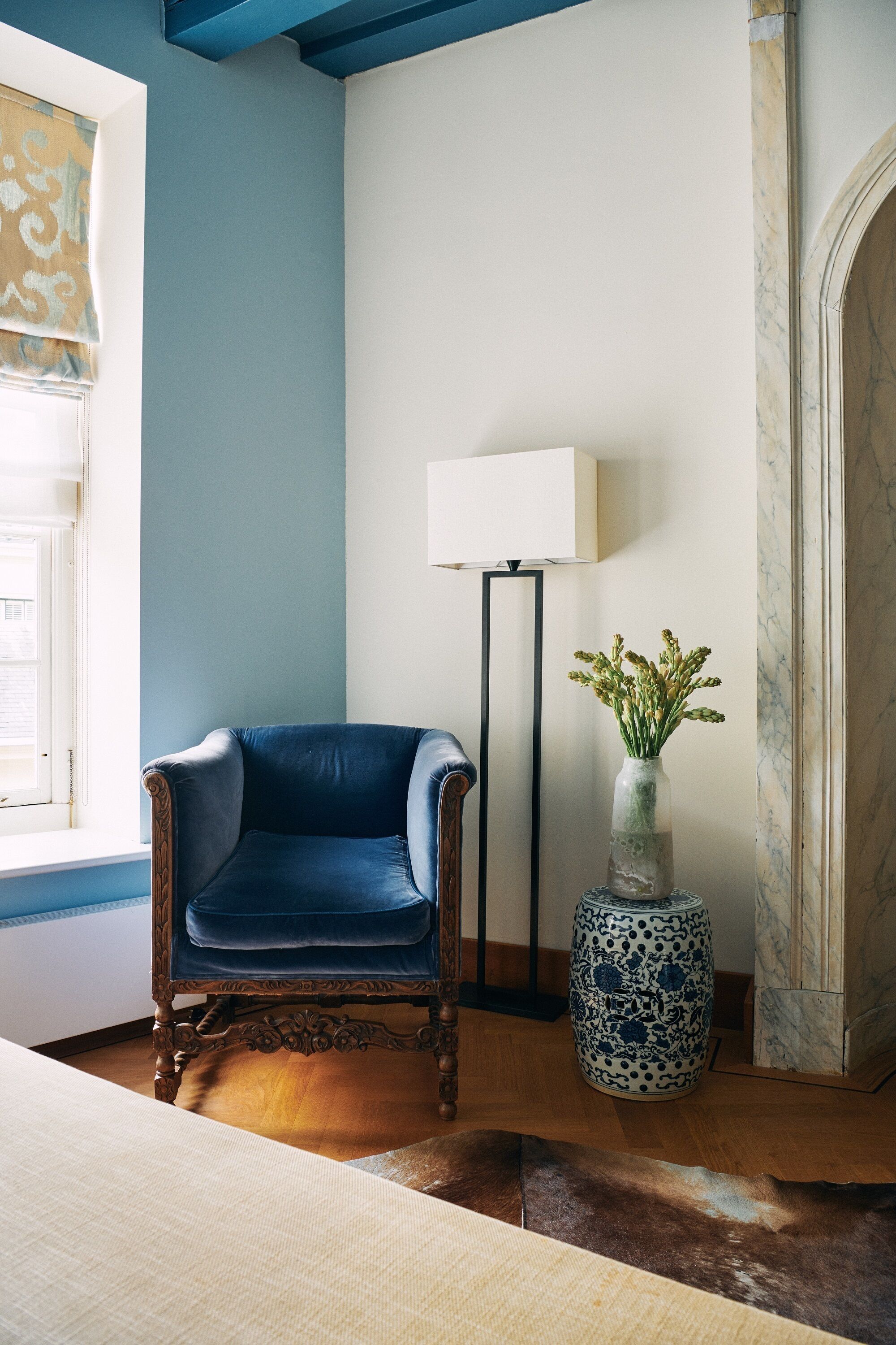 A room with a blue wall, a wooden chair, a floor lamp, and a vase with flowers.